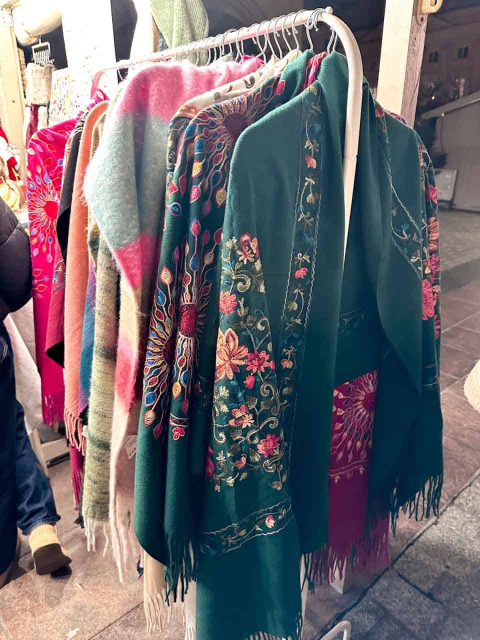 A rack of warm shawls and scarves in bold floral patterns, including an emerald green one with pink embroidery
