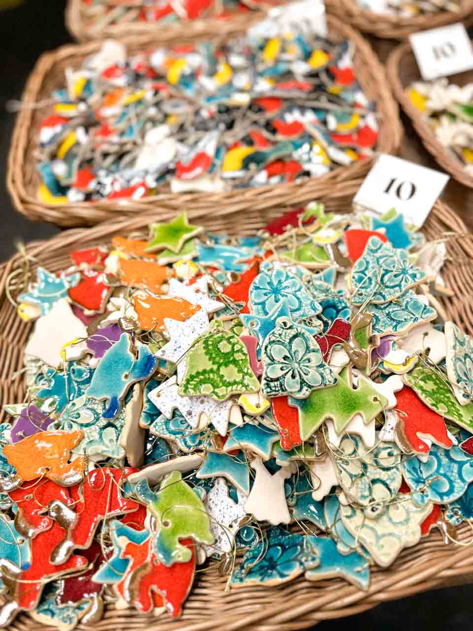 Ceramic Christmas tree ornaments in fun shapes and bright colours, laid out in wicker baskets marked with price tags