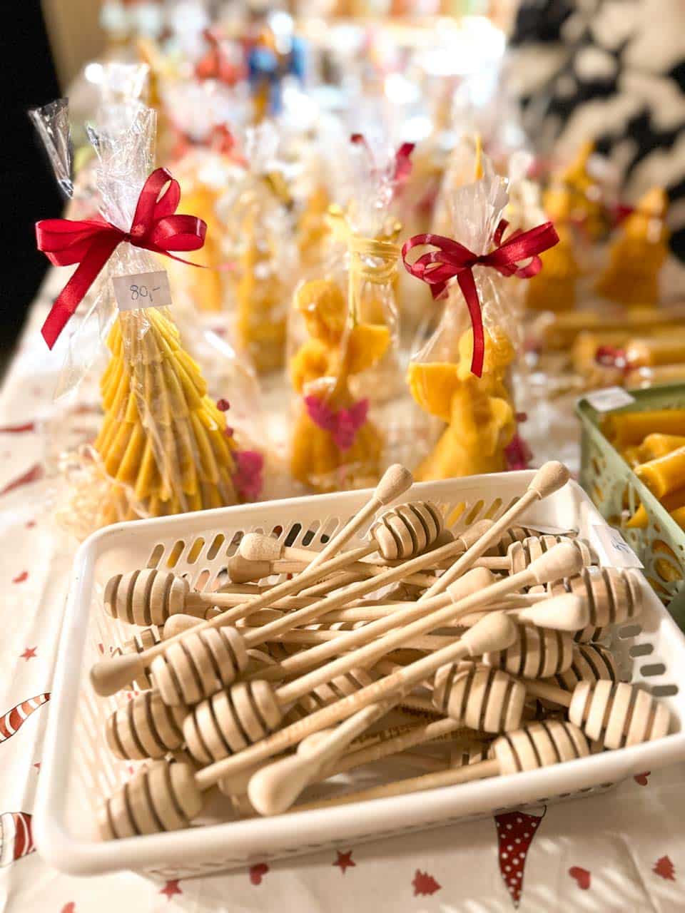 Beeswax candles shaped like angels and trees, wrapped in cellophane, with a tray of wooden honey dippers in front