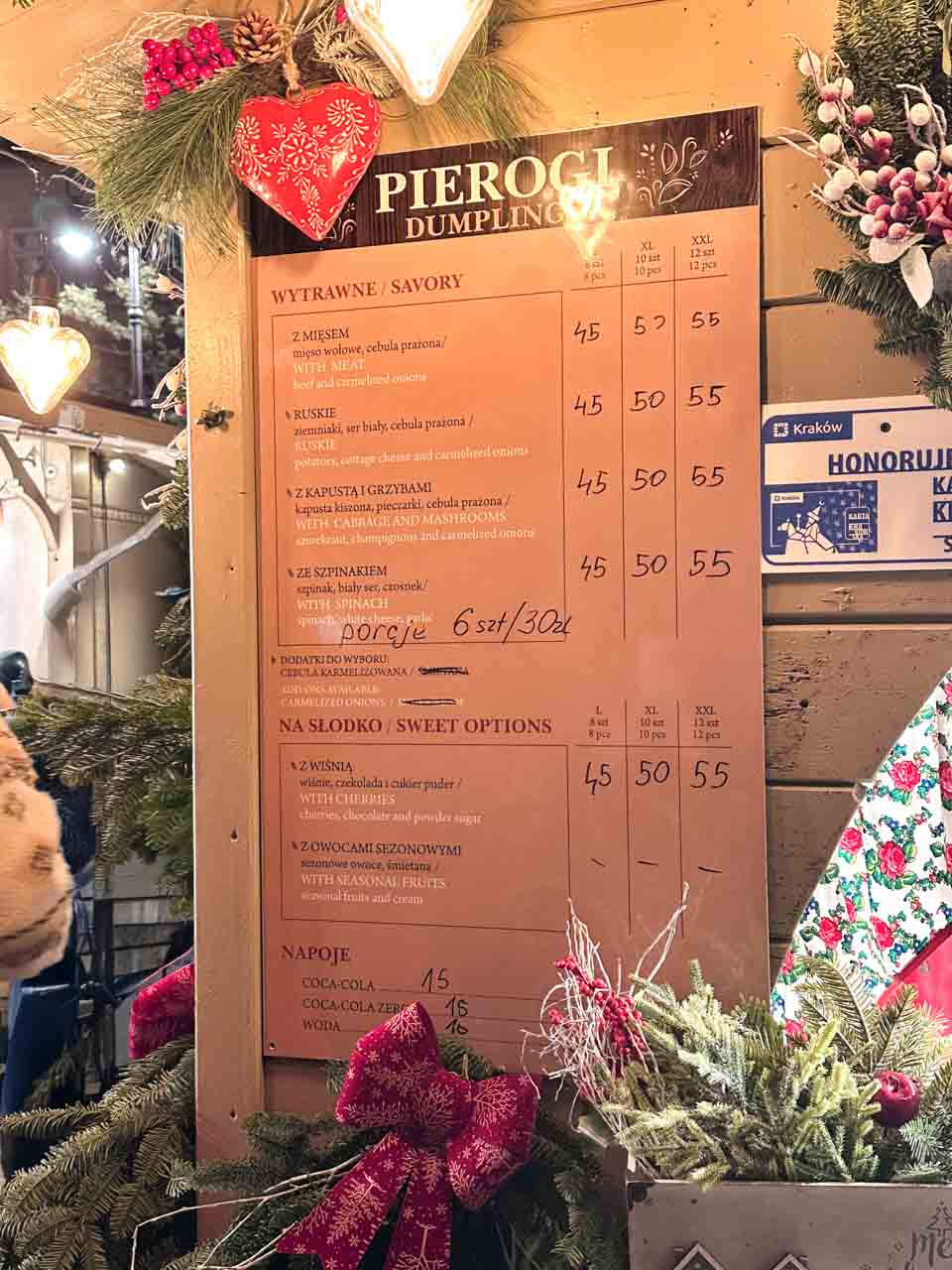 Festive pierogi menu board at the Kraków Christmas Market listing savoury and sweet dumplings in three portion sizes, decorated with pine branches