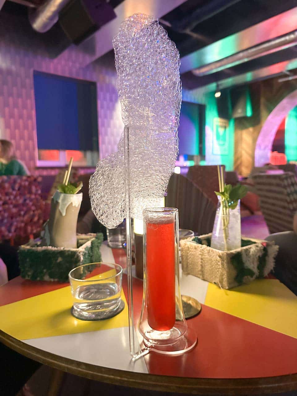 A tall glass of red cocktail with dramatic sugar foam, served on a colour-blocked table at MONK cocktail bar in Gdańsk, Poland