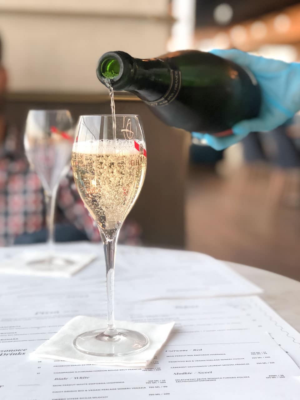 A glass of sparkling wine being poured at a stylish restaurant, placed on a menu-lined table with another glass in the background
