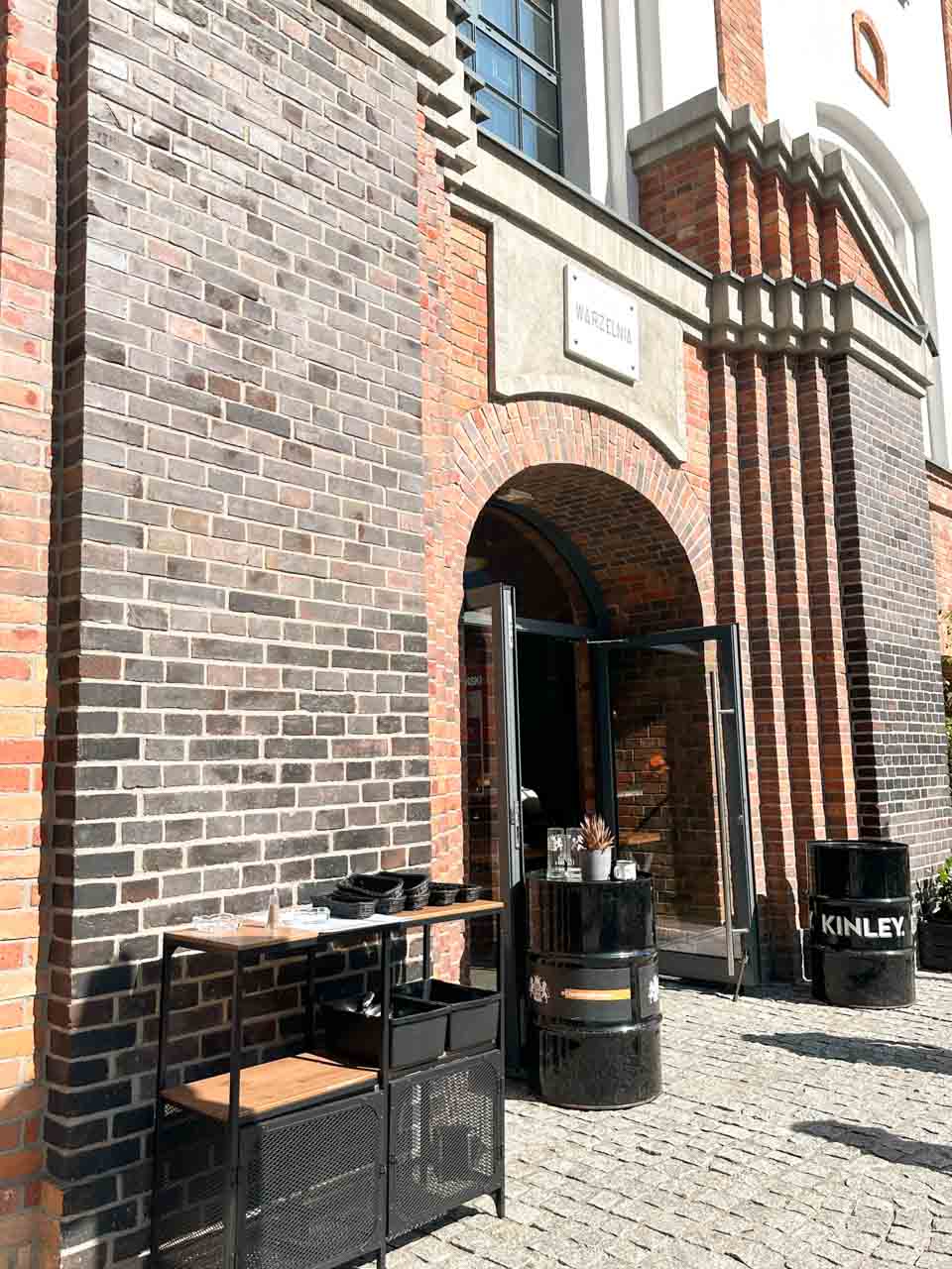 Entrance to the Nowy Browar Gdański brewery with red brick walls, a Warzelnia sign, and black Kinley-branded barrel tables