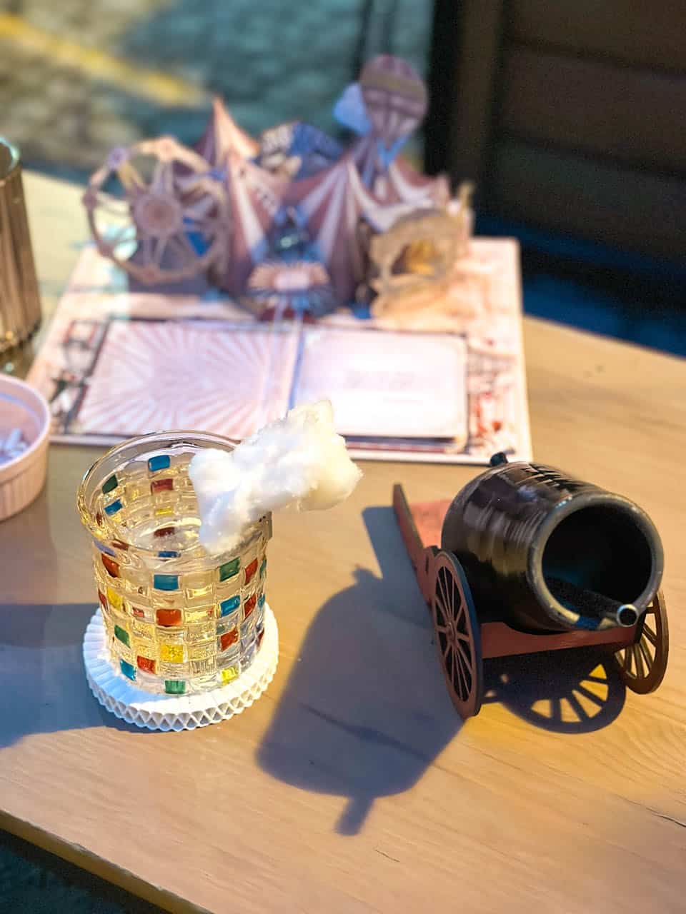 A cocktail topped with candy floss next to a miniature cannon on a table with a 3D pop-up menu at Flisak '76 in Gdańsk, Poland