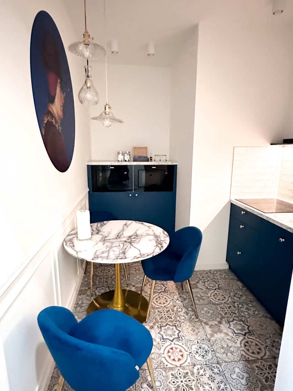 Small kitchen corner with a round marble table, blue velvet chairs, patterned tiles, and hanging glass lights
