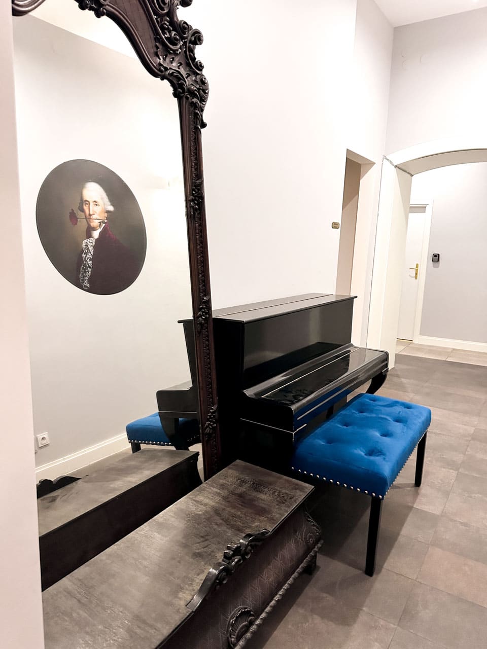 A hallway with a large ornate mirror, a piano, and a blue tufted bench next to a humorous portrait