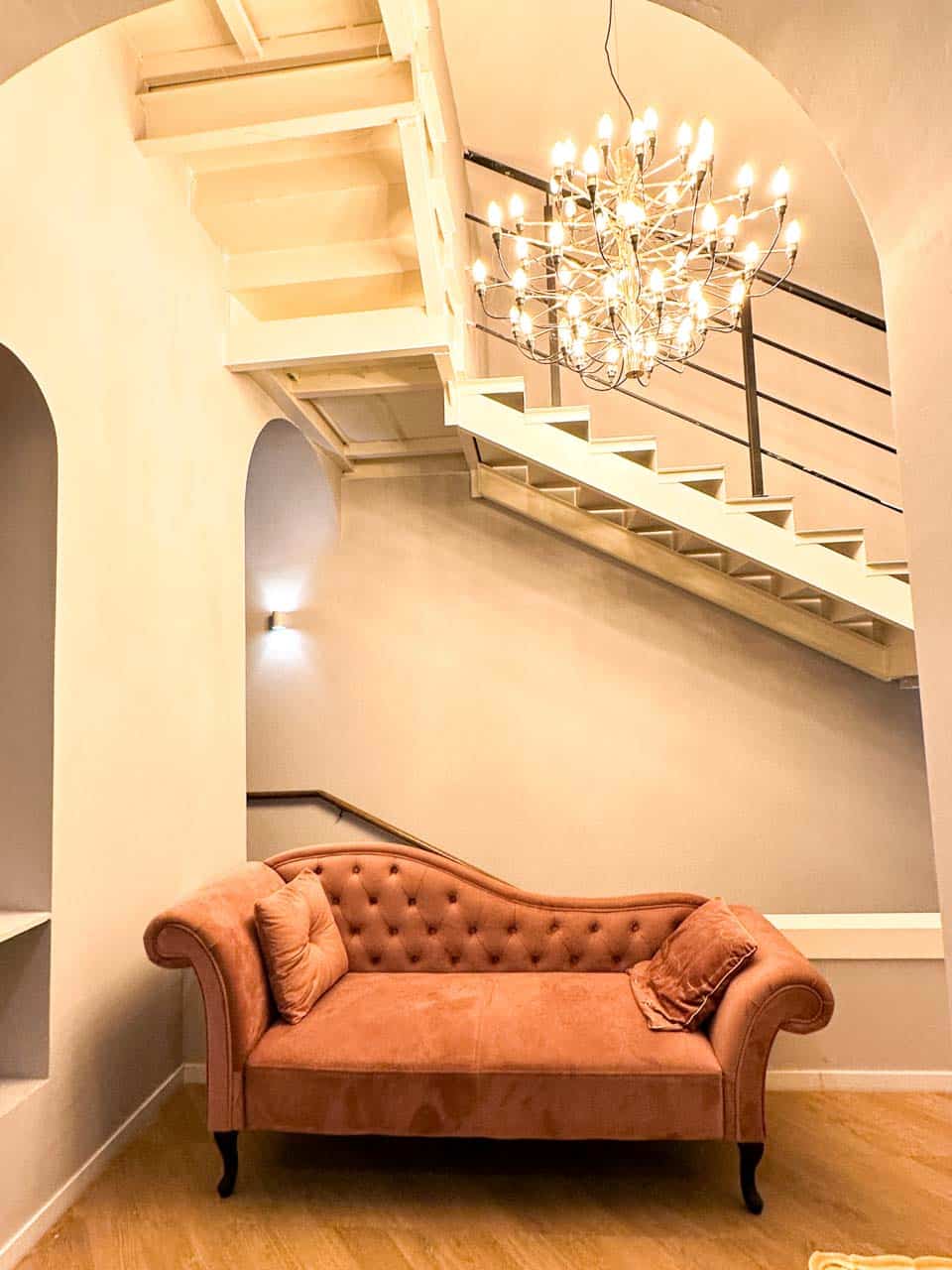 Peach-coloured vintage chaise lounge under a grand chandelier by a staircase with black railings and archways