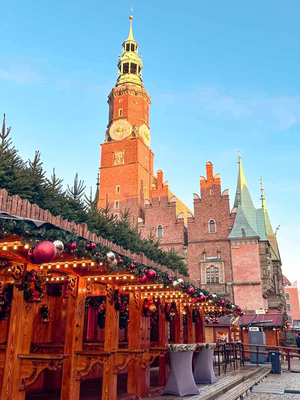 A view of the Wrocław Town Hall tower rising behind a festive wooden bar area lined with hanging baubles