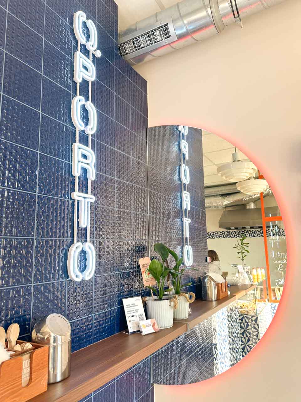 Blue tiled counter and a neon O.PORTO sign inside a small caf&eacute; with plants and a round mirror