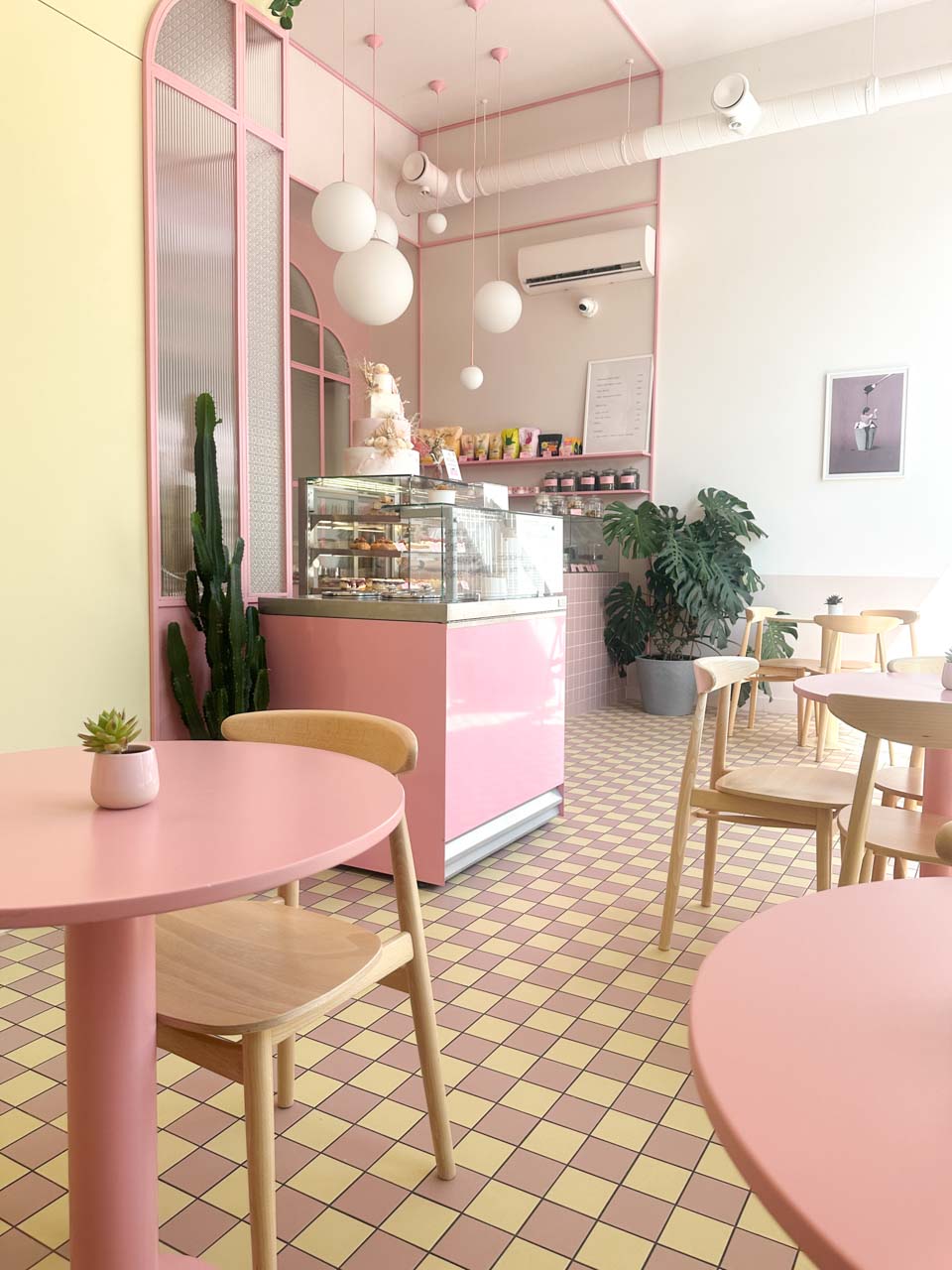 Pink caf&eacute; interior with tiled floor, round tables, plants, and a pastry counter