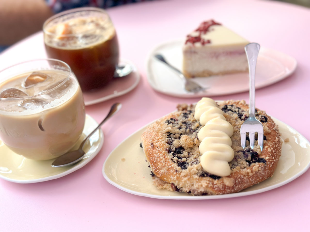 Bilberry crumble bun topped with vanilla cream beside iced coffees and a cheesecake slice on a pink table