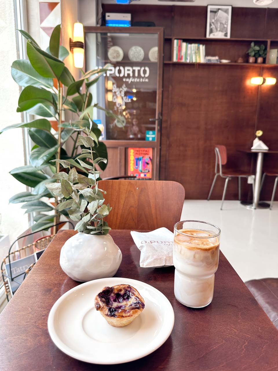 Iced latte and a pastel de nata on a wooden table beside a plant at O.PORTO Cafeteria in Gdańsk