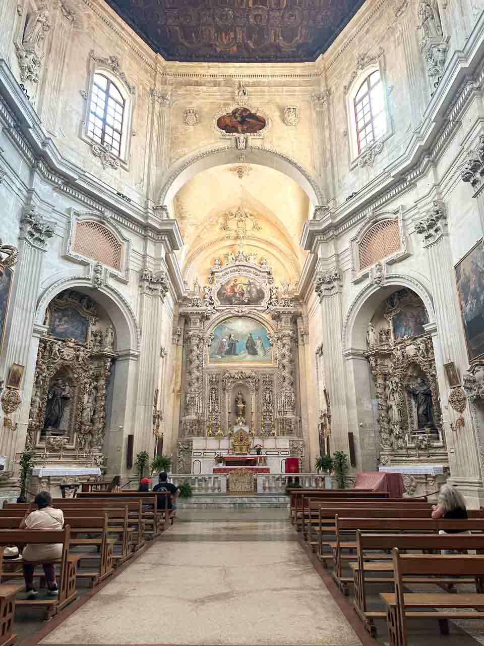 Interior of Chiesa di Santa Chiara in Lecce with ornate altars, wooden benches, and people sitting quietly