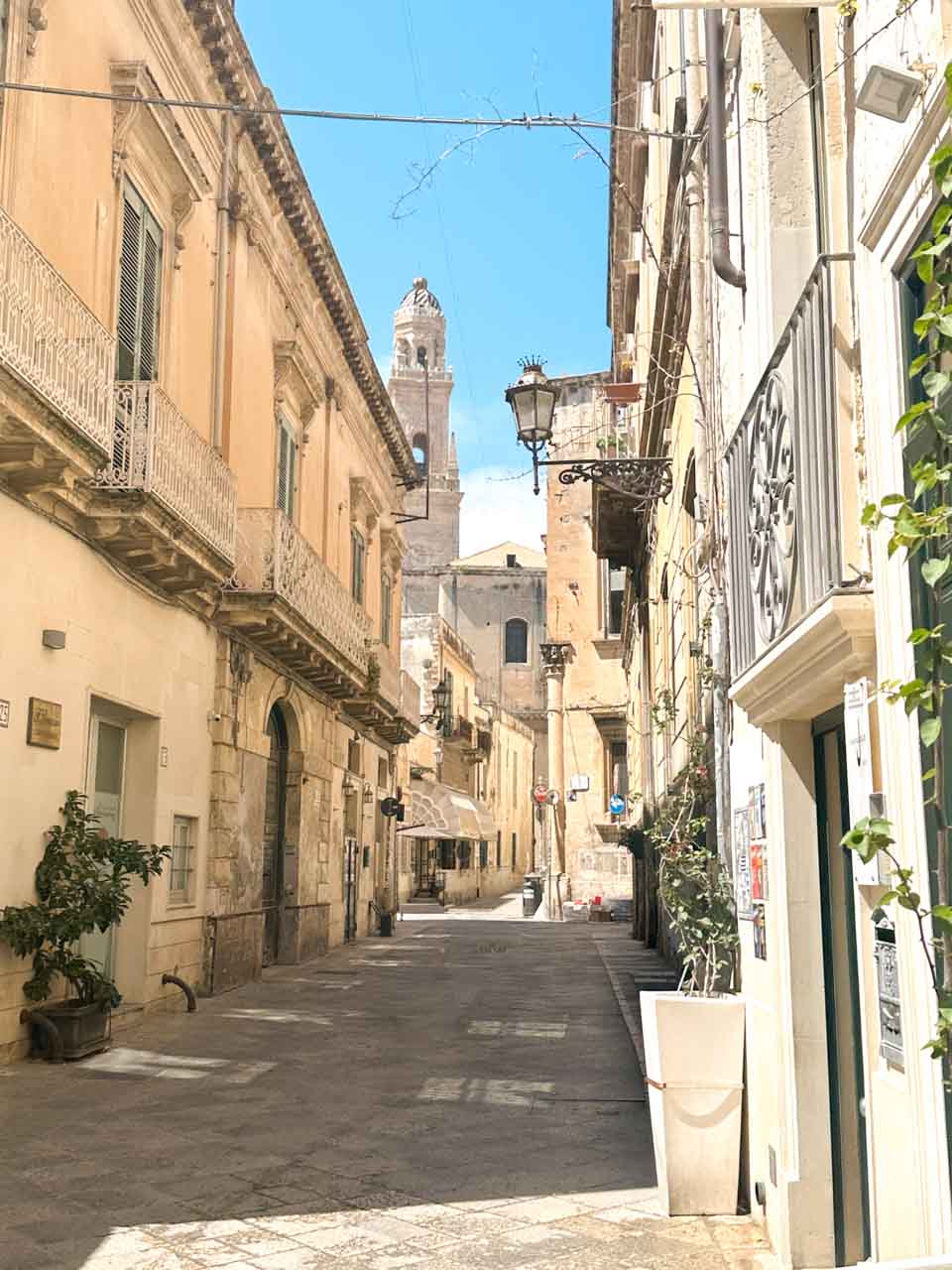 Sunlit street in Lecce&rsquo;s historic centre with balconies and the cathedral bell tower in the distance