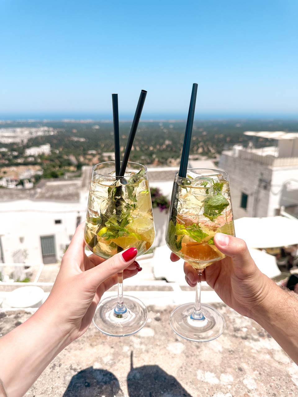 Two cocktails held up with a view over Ostuni&rsquo;s rooftops and countryside in the distance