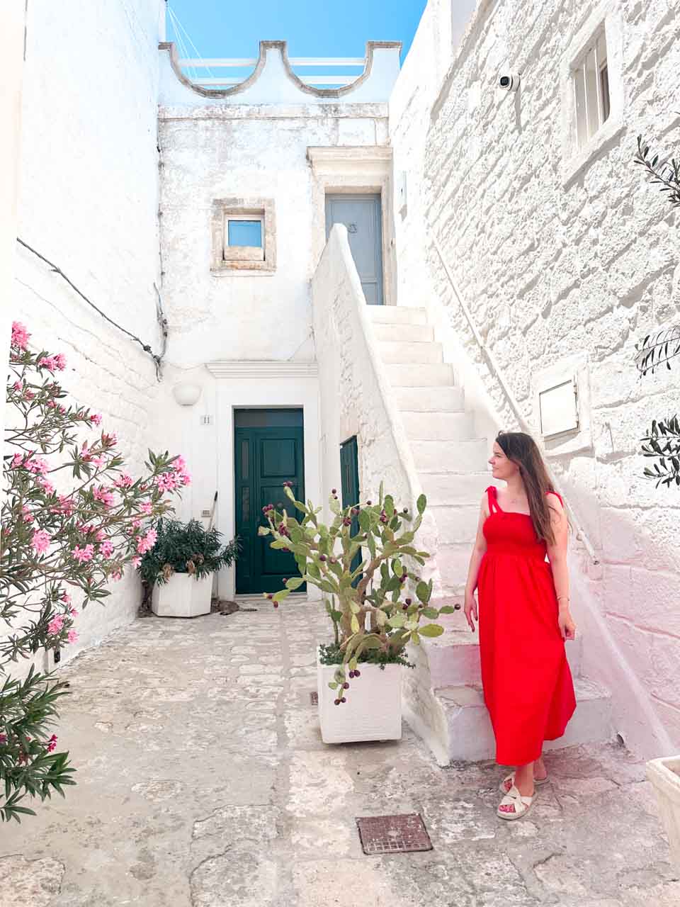 Woman in a red dress standing by a staircase and a potted cactus in a whitewashed courtyard in Ostuni, Italy