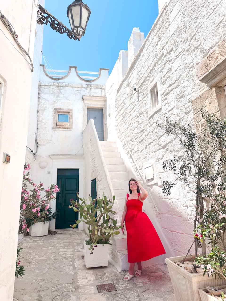 Woman in a red dress posing in a whitewashed courtyard with plants and stone steps in Ostuni, Italy