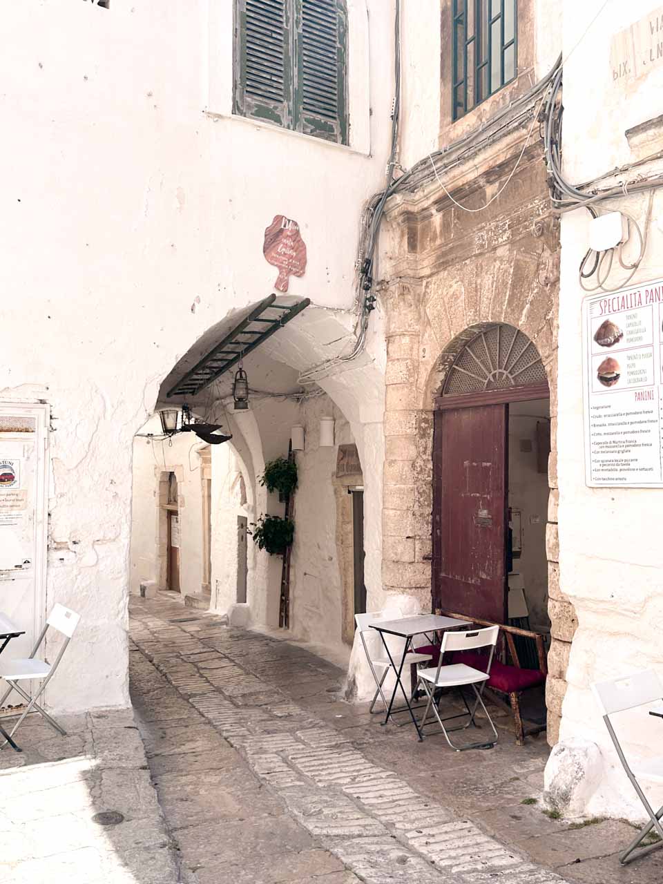Small caf&eacute; table set along a quiet white alley with an arched doorway in Ostuni, Italy