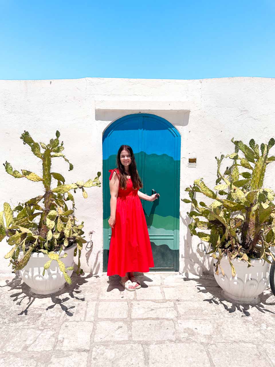 Woman in a red dress standing by a blue and green door with cactus plants in Ostuni
