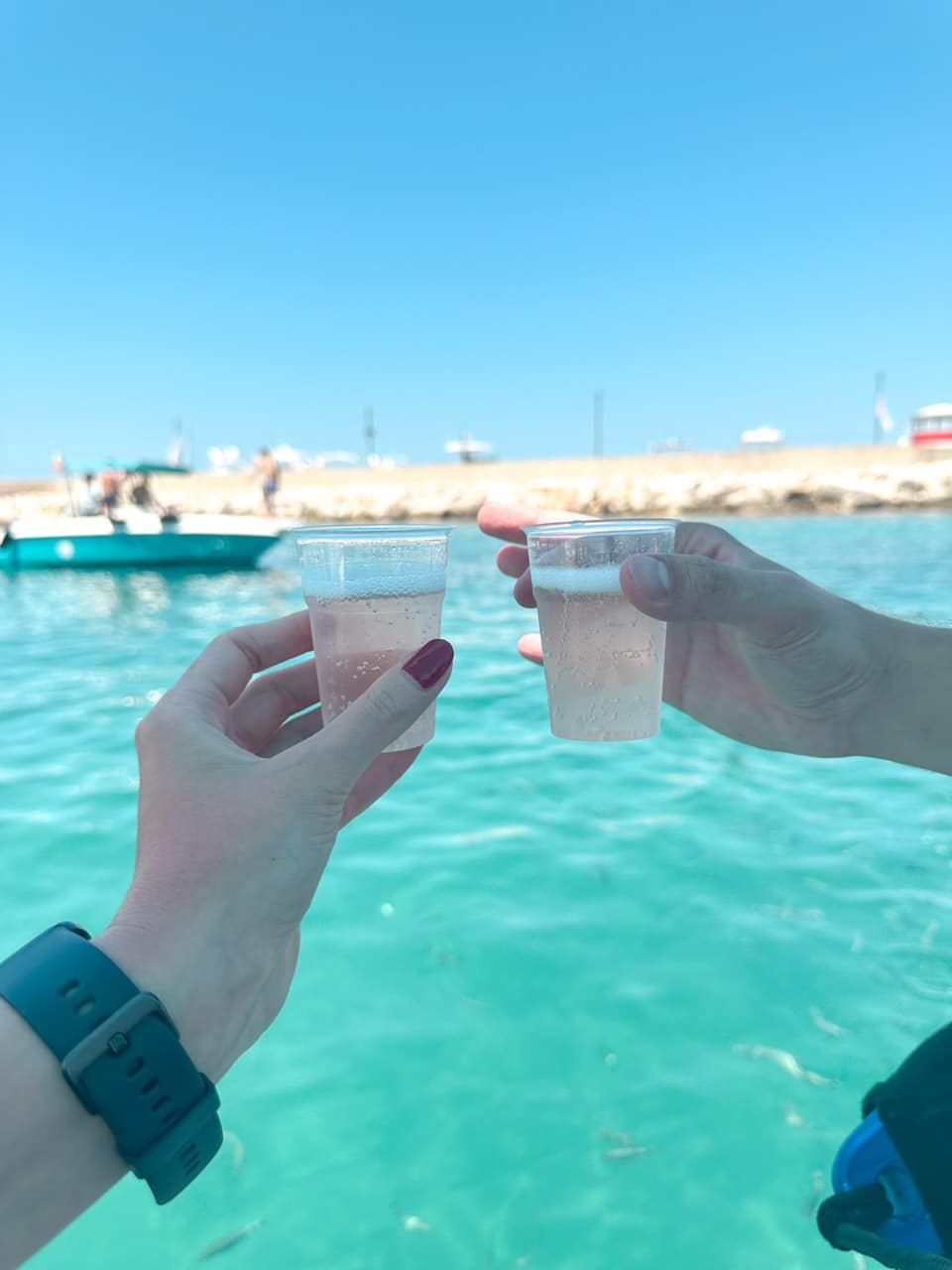 Two plastic cups of sparkling wine held up over the sea near the harbour in Polignano a Mare, Italy