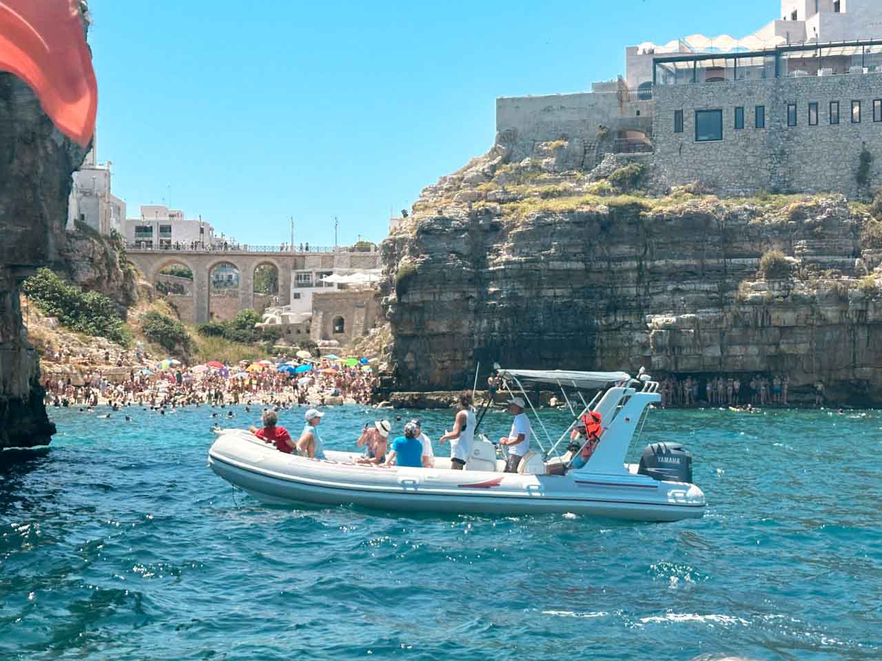 A boat near the Lama Monachile beach with a stone bridge and crowds on the shore in Polignano a Mare, Italy