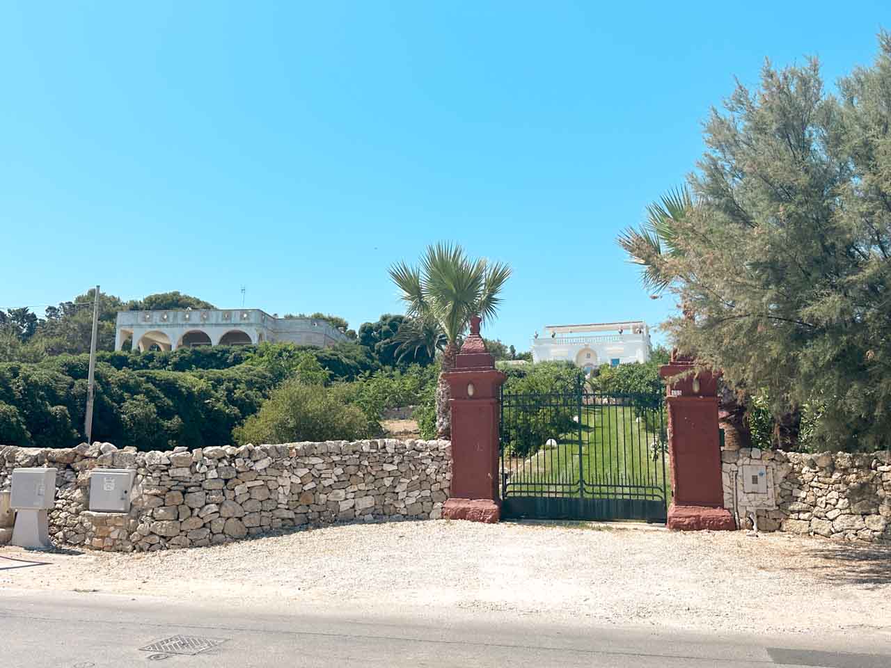 A gated entrance with palm trees and a white villa set back along a quiet road in Polignano a Mare, Italy