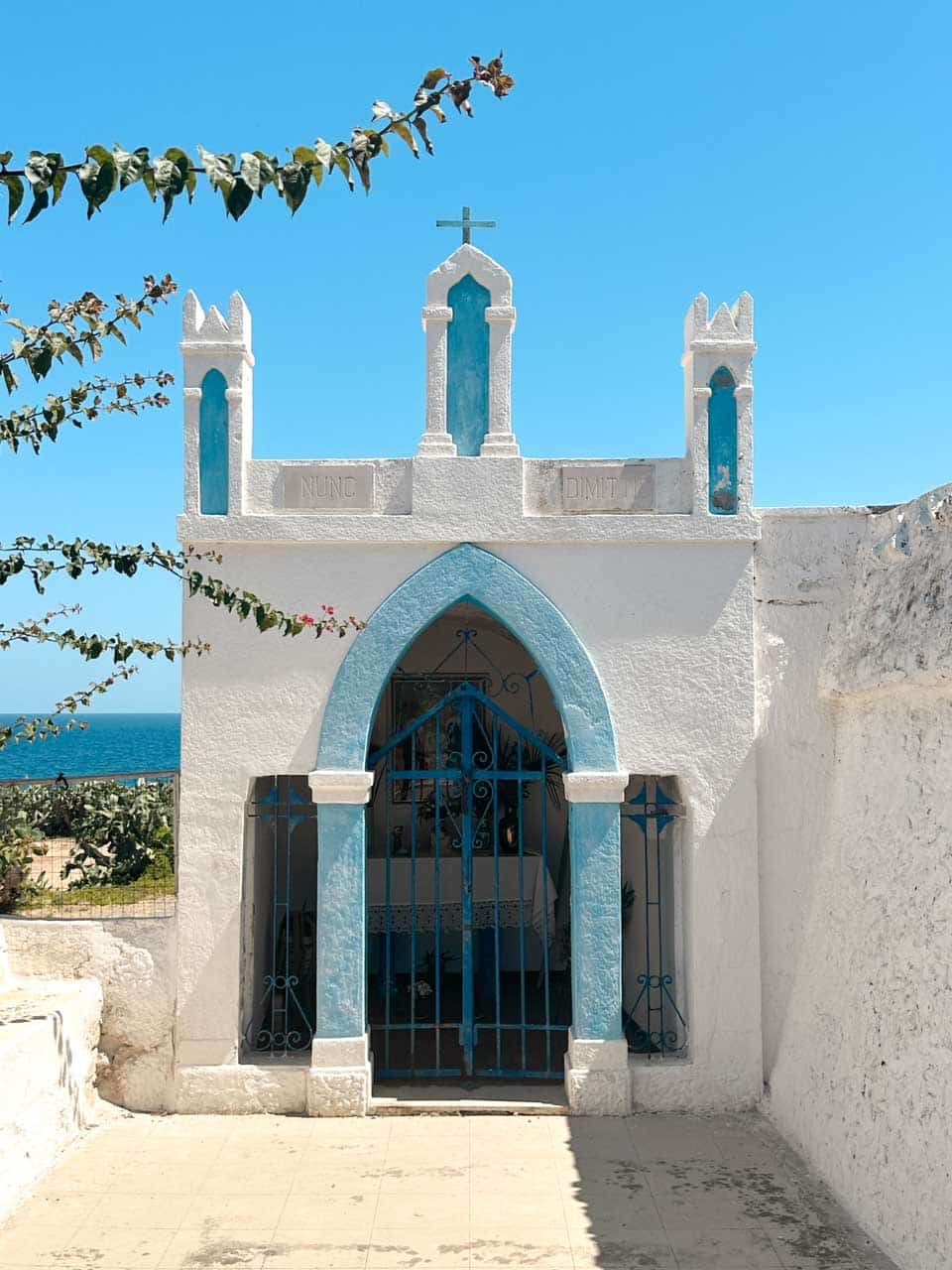 A small white chapel with blue details and a cross overlooking the sea in Polignano a Mare, Italy