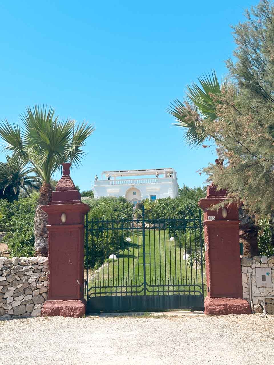 A gated entrance with red pillars and a white villa surrounded by greenery in Polignano a Mare, Italy
