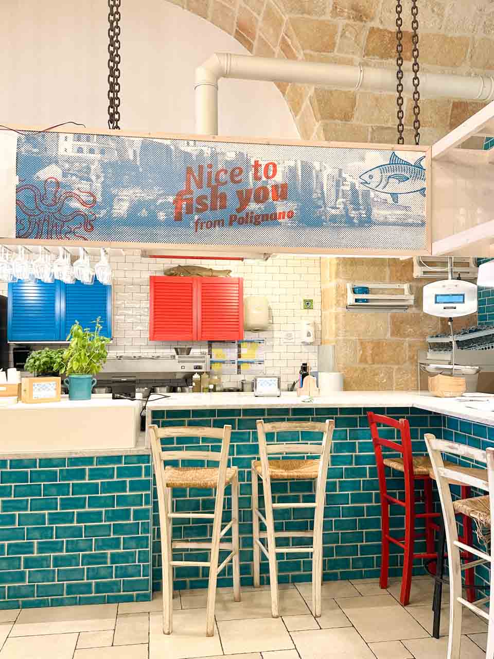 Bright interior of Pescaria in Polignano a Mare with turquoise tiles, wooden stools, and a &ldquo;Nice to fish you from Polignano&rdquo; sign