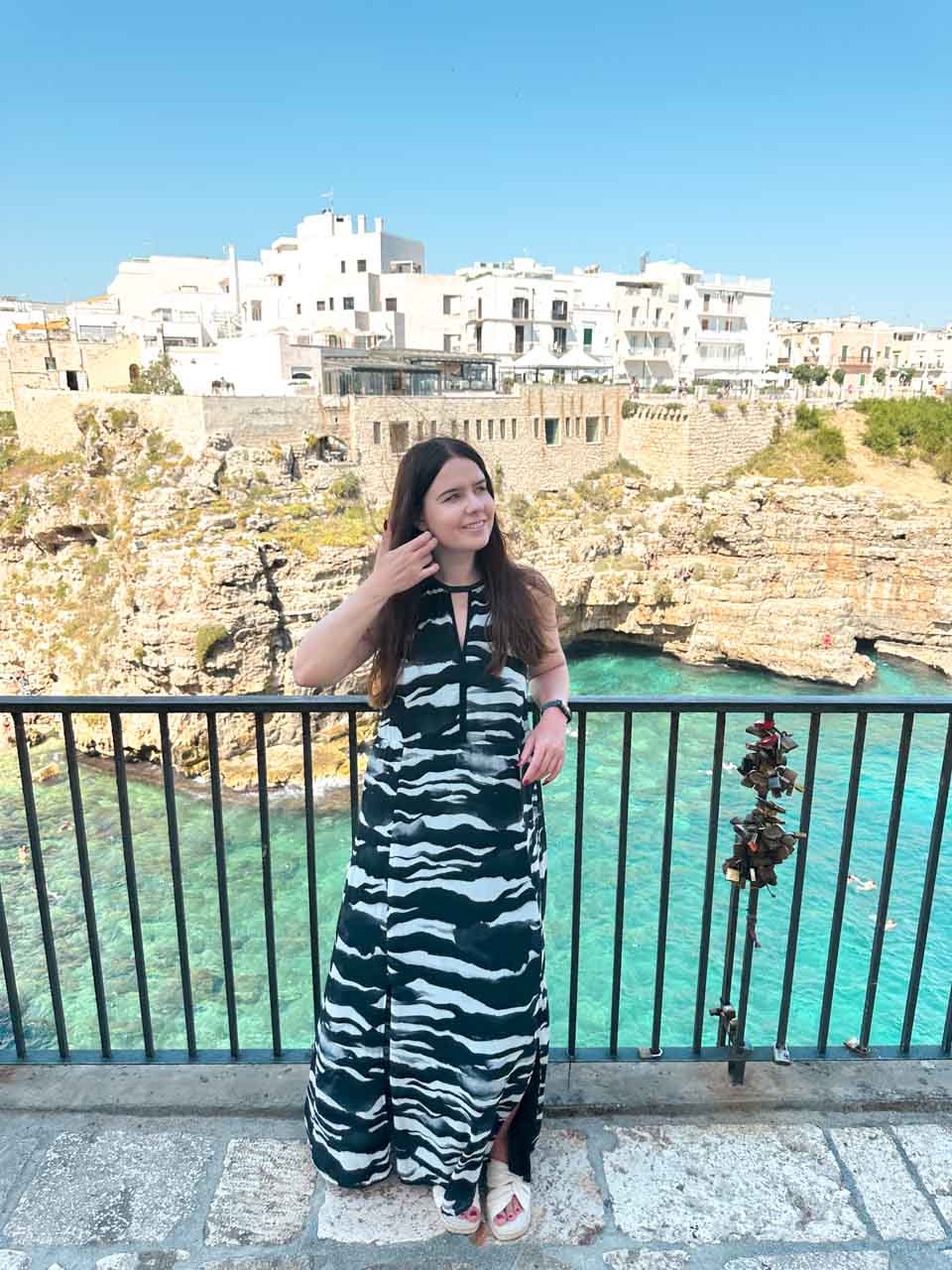 Woman in a black and white maxi dress standing by a railing overlooking the turquoise sea and cliffs in Polignano a Mare