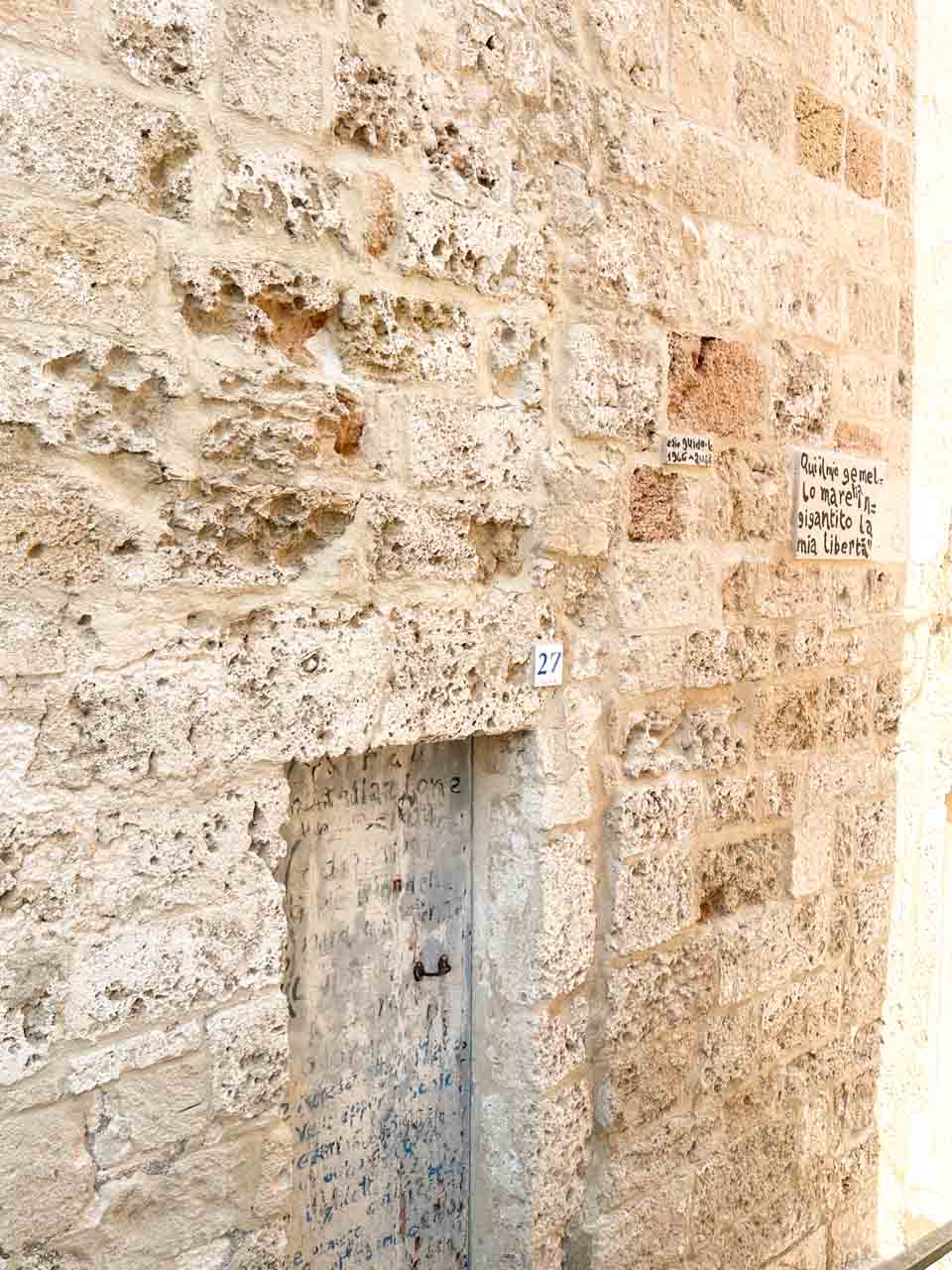 Weathered stone wall with a small door, house number, and handwritten Italian quotes in Polignano a Mare