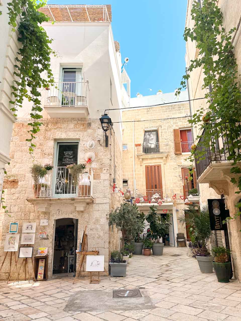Small courtyard in Polignano a Mare with stone buildings, balconies, plants, and an art shop
