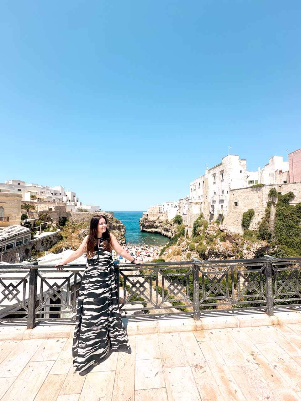 A woman in a black and white maxi dress at a viewpoint overlooking the Lama Monachile beach and cliffs in Polignano a Mare, Italy