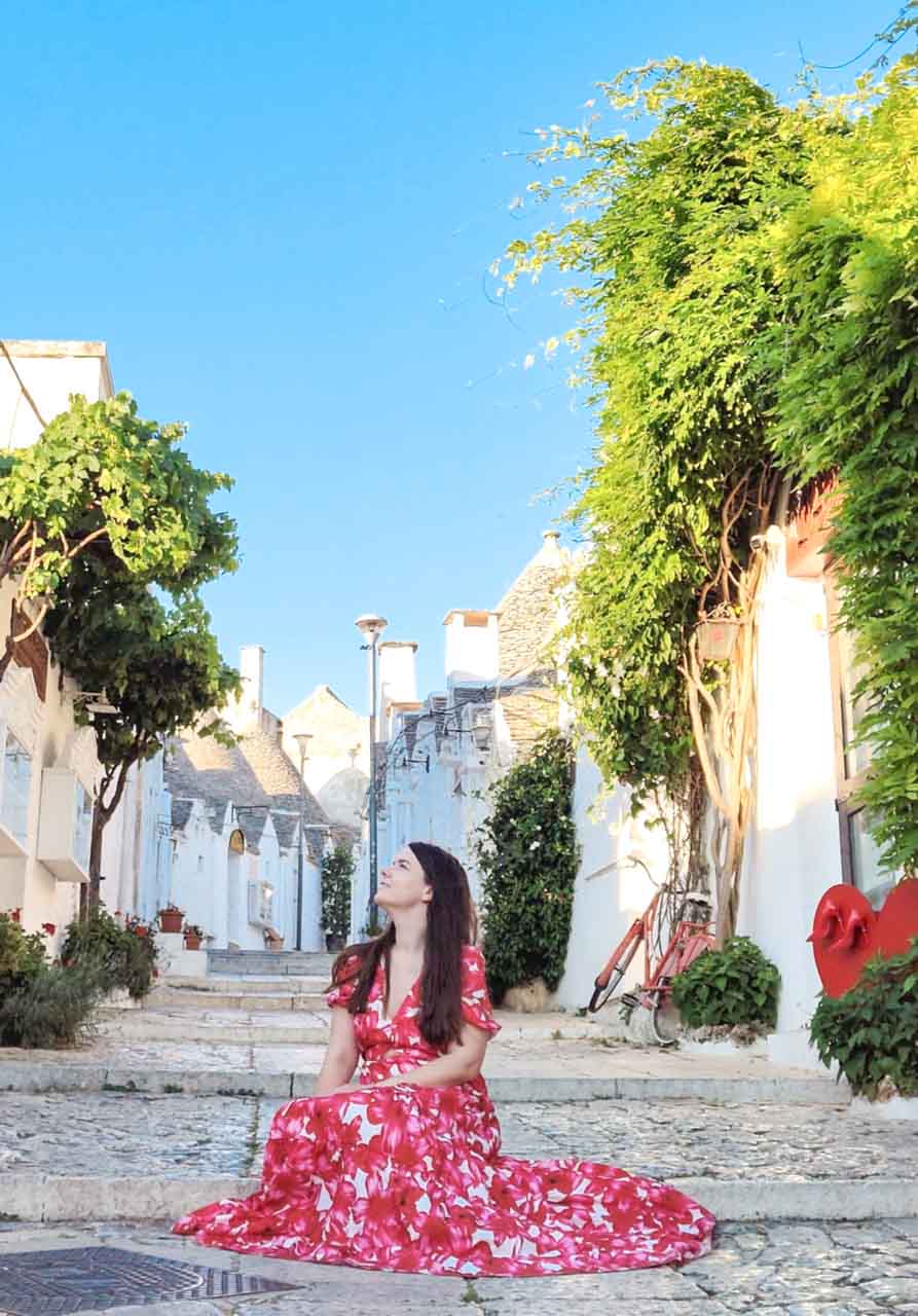 Woman in a red, pink, and white dress sitting on stone steps between white trulli houses in Alberobello, Italy