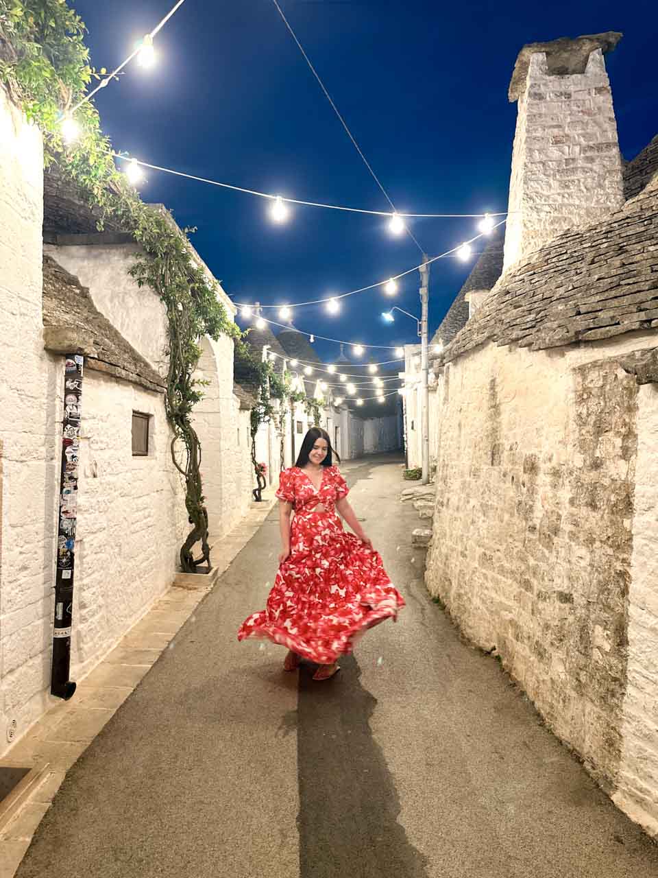 Woman in a pink, red, and white dress twirling beneath string lights on a trulli-lined street in Alberobello at night