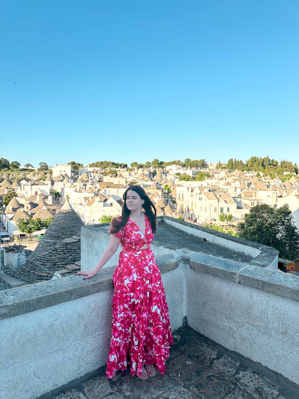 Woman in a red, pink, and white dress standing on a terrace overlooking trulli rooftops in Alberobello, Italy
