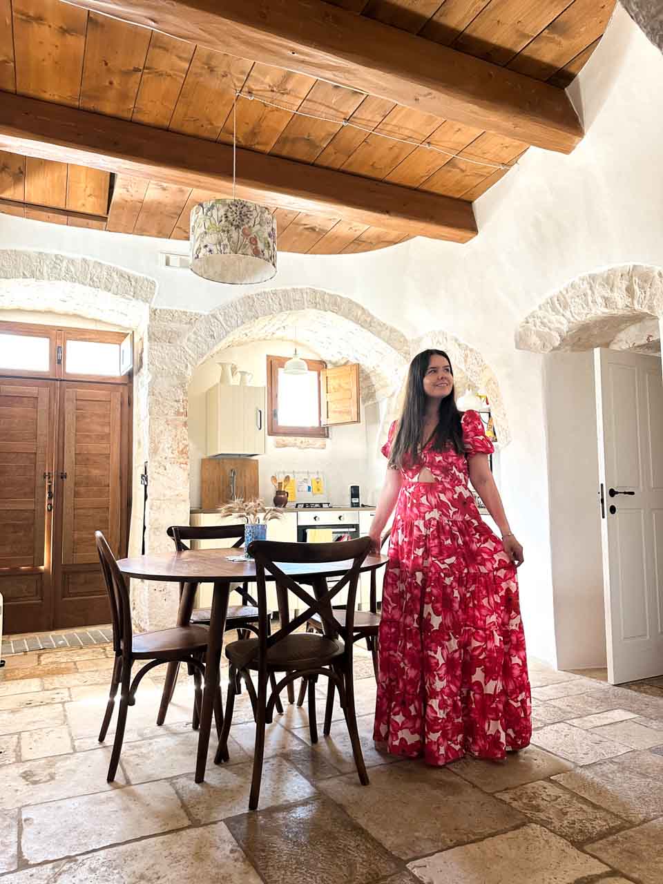 Woman in a red, pink, and white dress standing inside a rustic trullo house with stone walls and a wooden ceiling