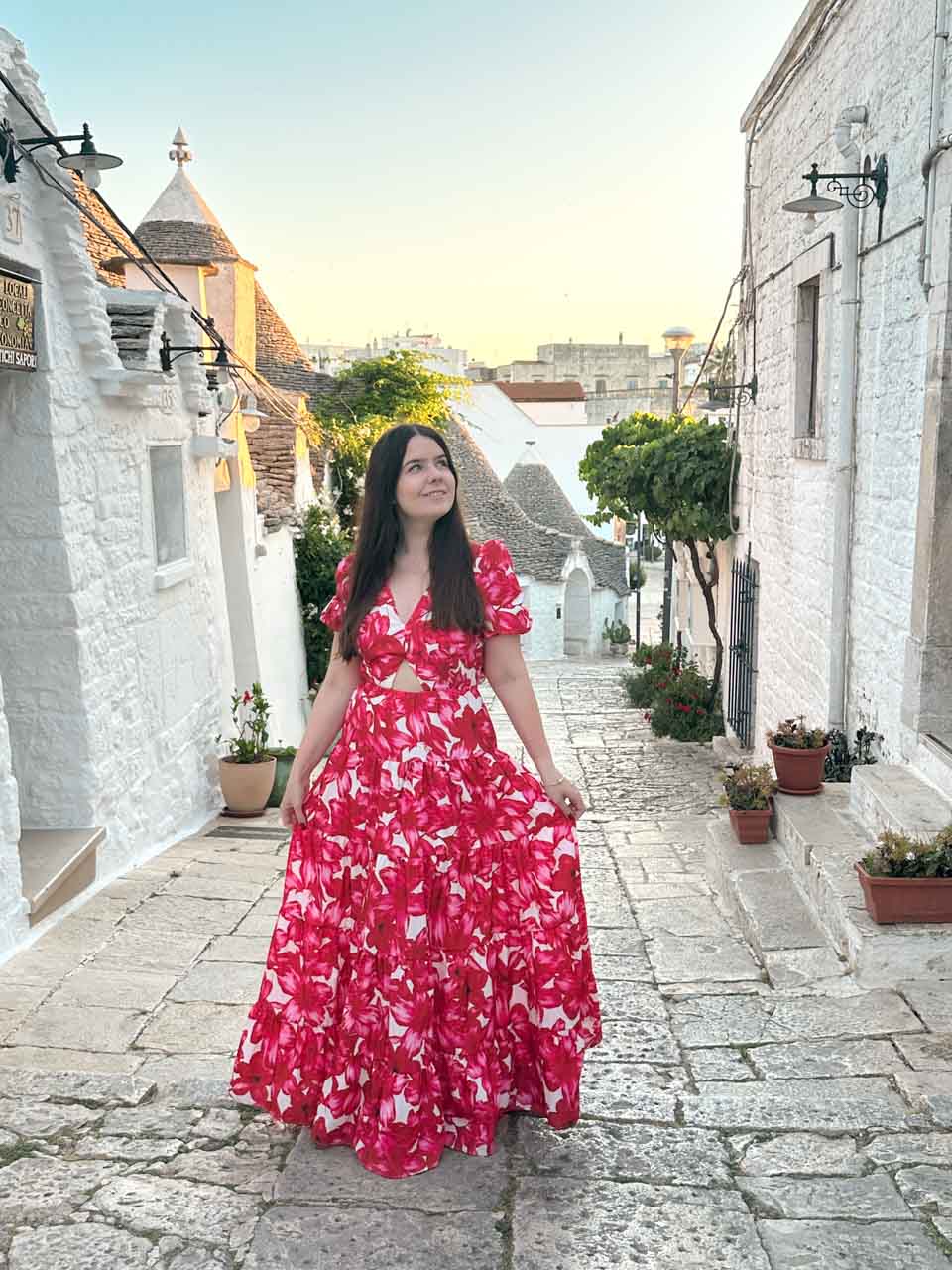 Woman in a red, pink, and white dress standing on a stone street with trulli behind in Alberobello, Italy