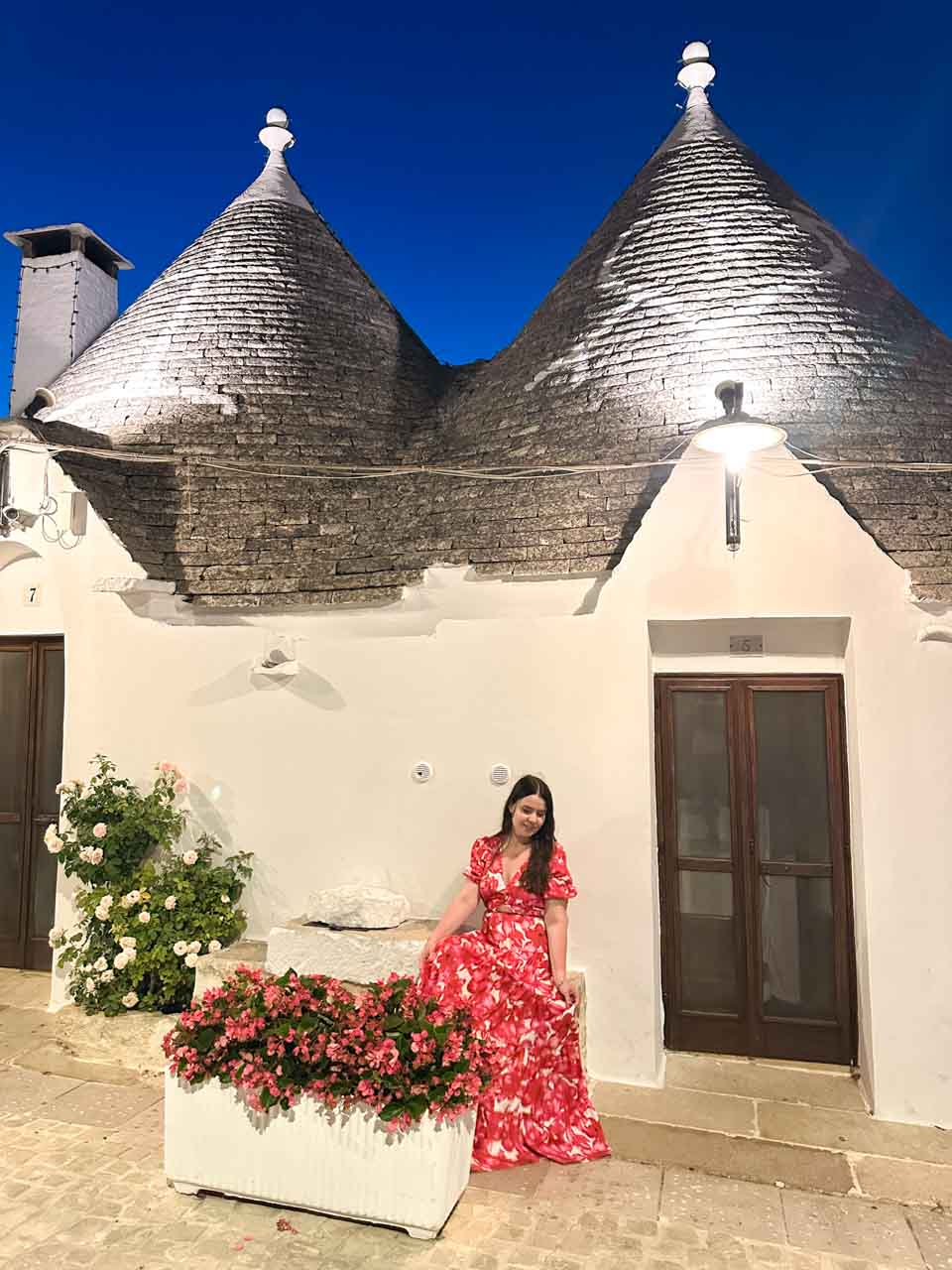 Woman in a red, pink, and white dress sitting by flowers outside a trullo house at night