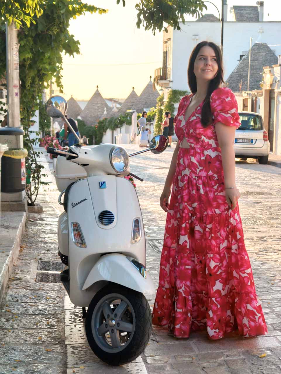 Woman in a red, pink, and white dress standing next to a white Vespa on a street lined with trulli houses