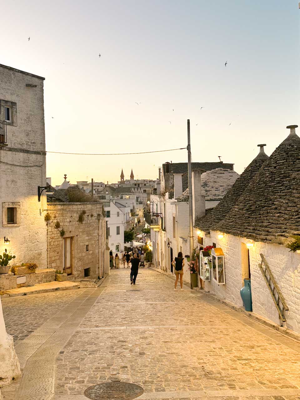Sloping street in Alberobello at sunset with trulli roofs and people walking