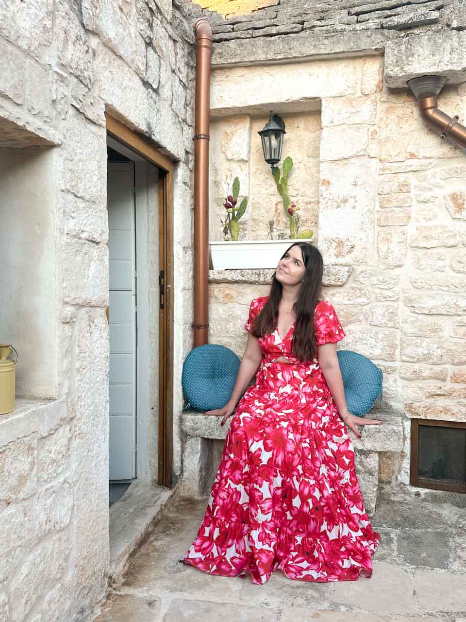 Woman in a red, pink, and white dress sitting outside a stone trullo in Alberobello, Italy