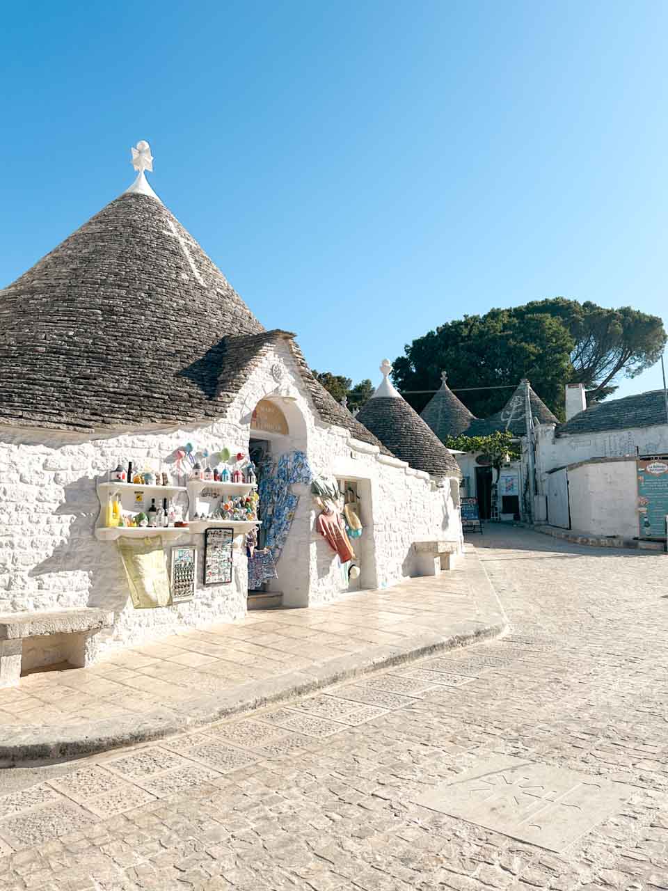 White trullo house with a souvenir display along a quiet sunlit street in Alberobello, Italy