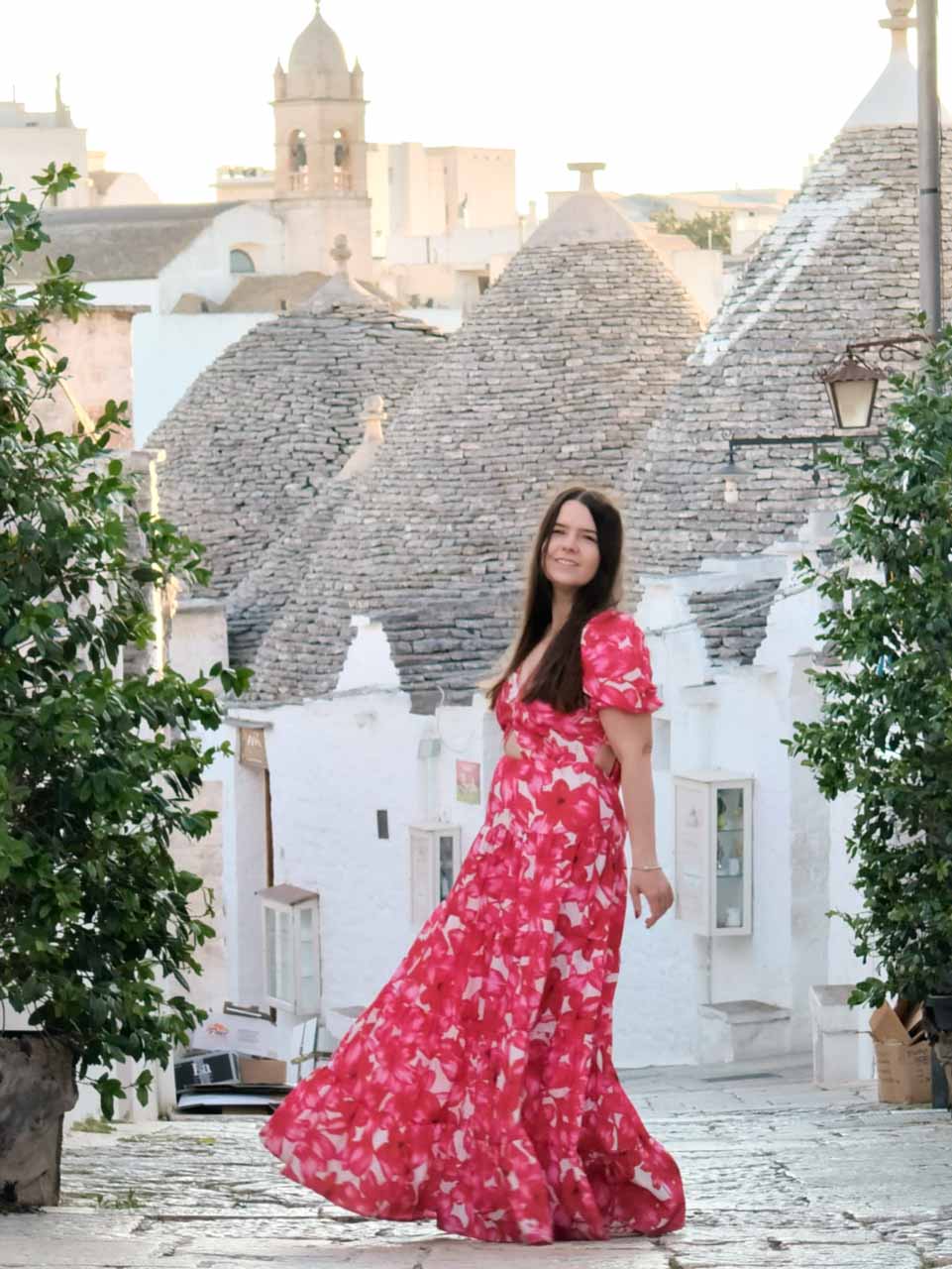 Woman in a red, pink, and white dress posing with trulli rooftops in the background in Alberobello