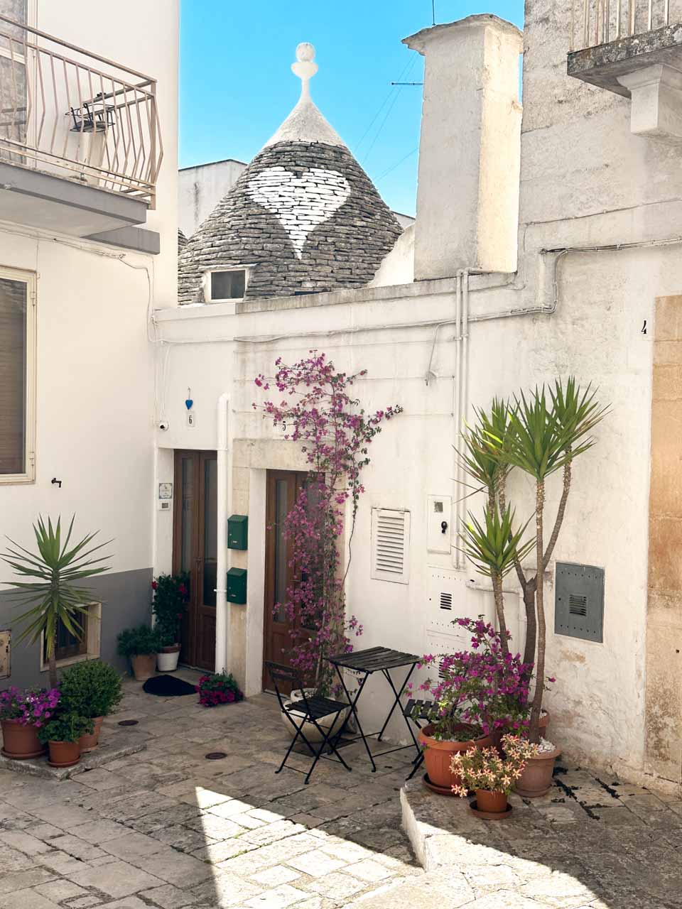 Small courtyard with flowering plants and a trullo roof painted with a white heart in Alberobello