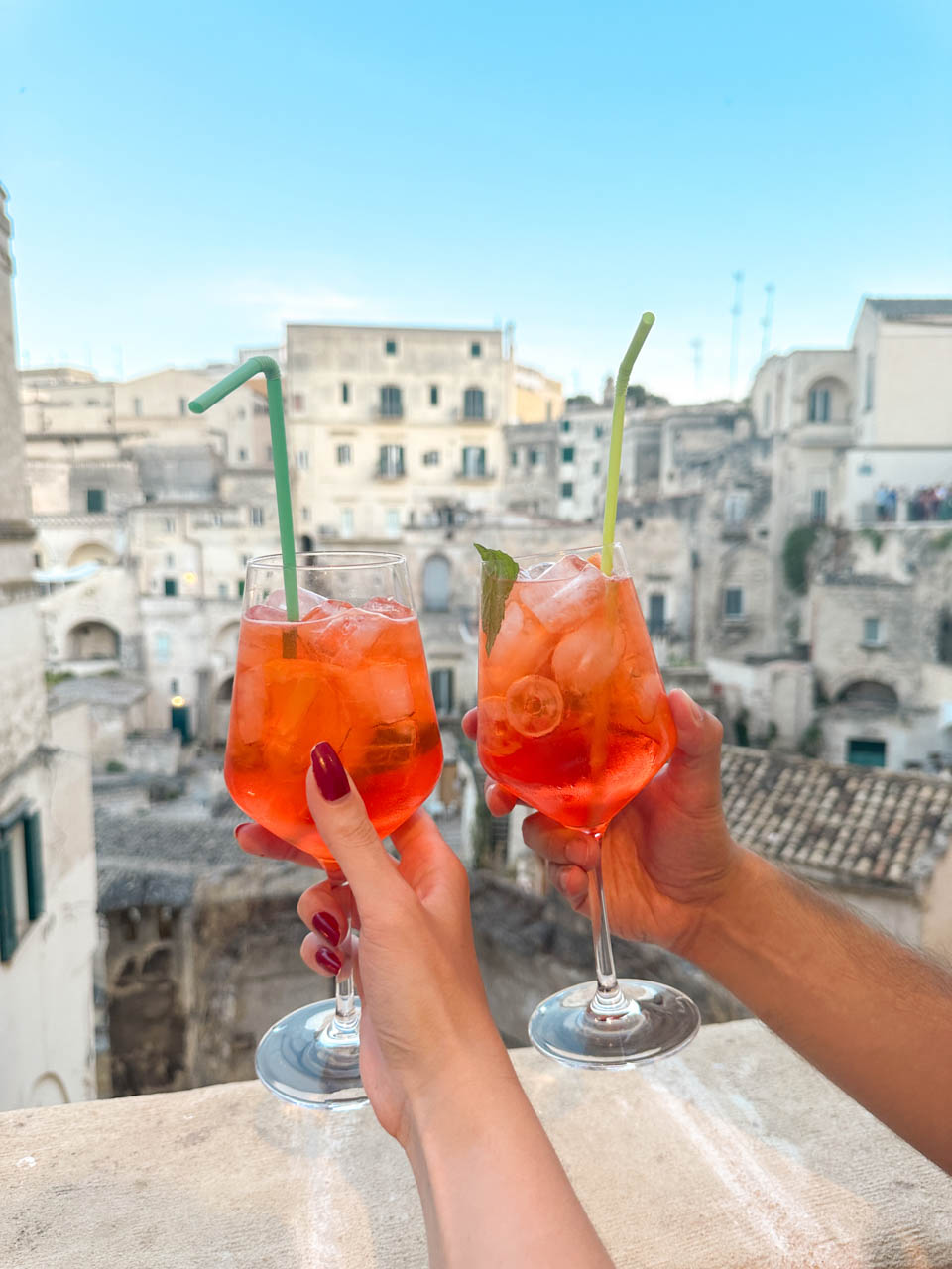 Two people holding Aperol Spritz cocktails overlooking the Sassi district of Matera