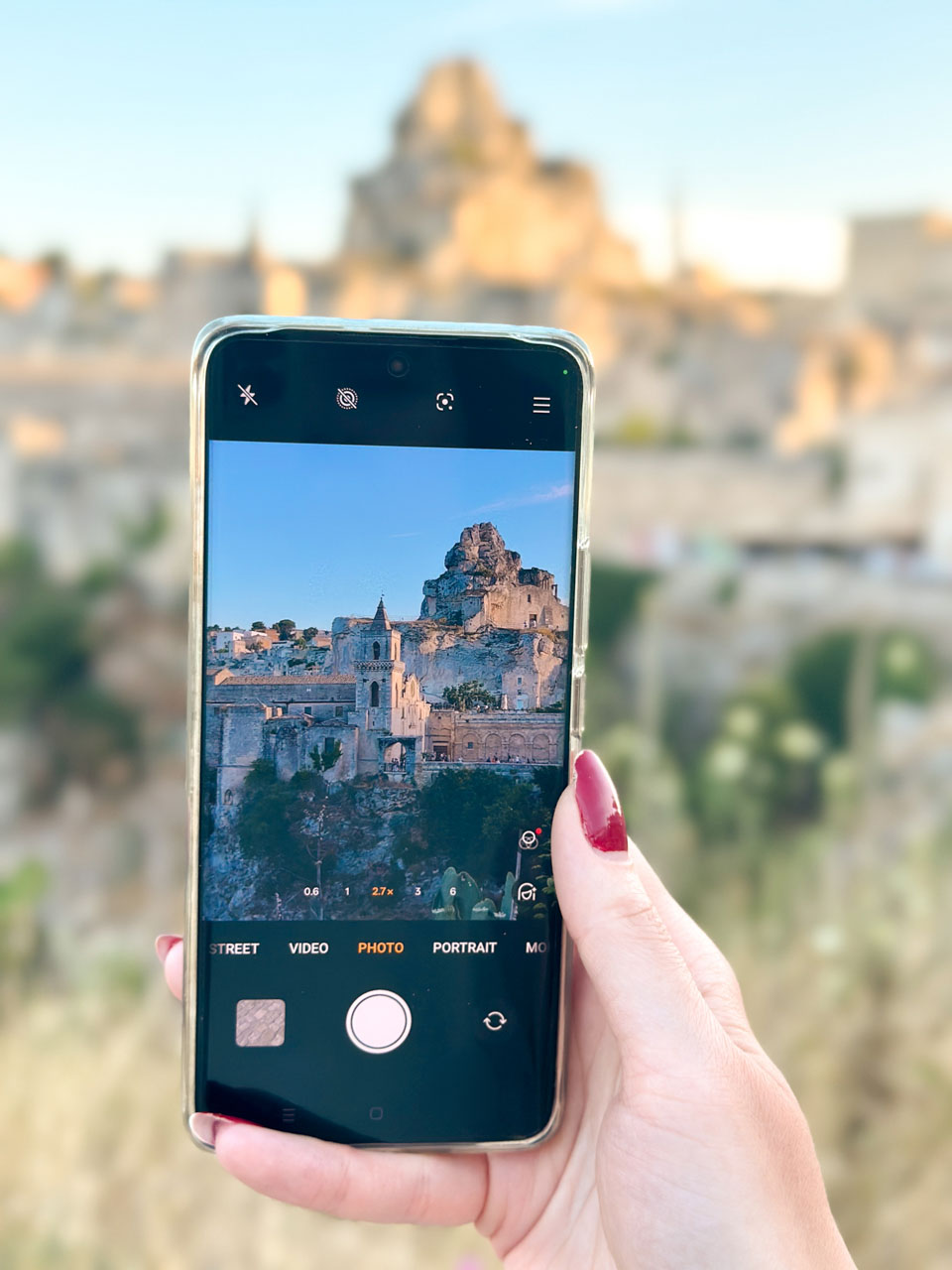 Close-up of a woman's hand holding a smartphone photographing Matera's Sassi at sunset