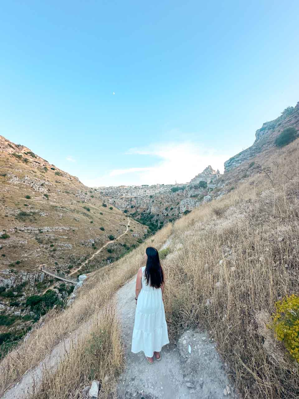 A woman in a white maxi dress standing on a hillside path overlooking the Gravina canyon and Matera in the distance