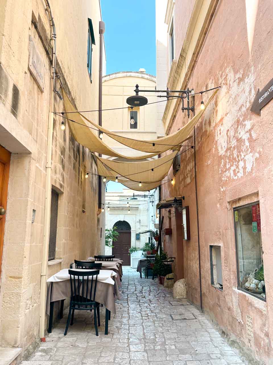 Outdoor restaurant tables beneath shade sails in a narrow street in Matera's historic centre