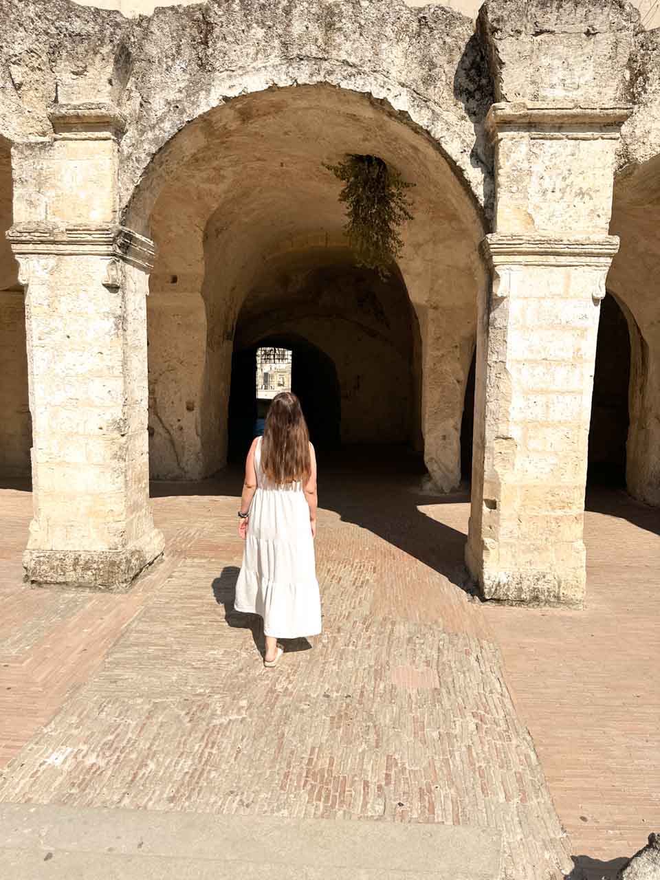 A woman in a white maxi dress walking through the arched passage leading to the lower level of the Tre Archi viewpoint in Matera